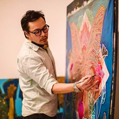 Image of a man with dark hair and glasses wearing a white long-sleeved shirt painting on a very large, colorful canvas