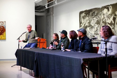 Panel discussion in a modern gallery space, with six speakers seated at a long table and one moderator, set against large abstract artwork and colorful paintings on the walls.