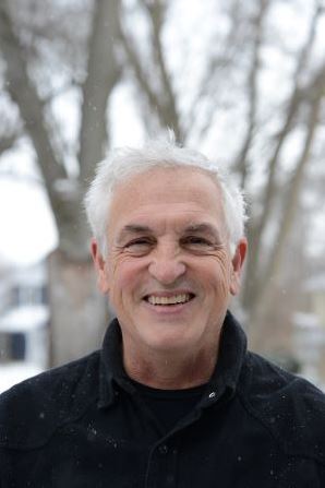 Adult with white hair smiling outdoors in a snowy winter setting, wearing a dark jacket lightly dusted with snowflakes; leafless trees and snow-covered ground in the background. 