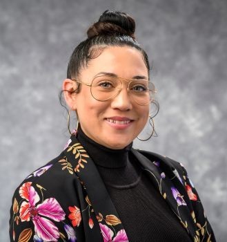Professional headshot of a smiling person with long dark hair, wearing hoop earrings, a black blazer, and a dark purple top against a dark gray background.