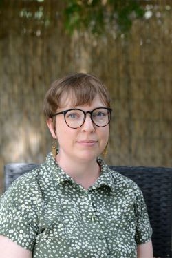 Headshot photograph of a woman with short light brown hair, black circular glasses, gold earrings, and a light green top with white spots. 