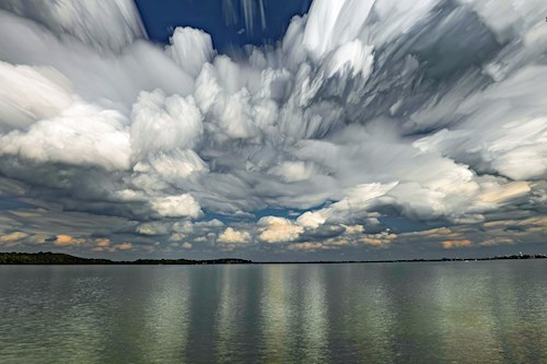 Landscape photograph of a glassy lake with thick, billowing white clouds above. The clouds have blurred as the photo compiles multiple shots over time.