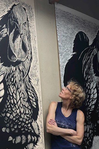 Image of woman with short blonde curly hair wearing glasses and a blue tank top looking up at a woodblock print of a chicken that is twice her height