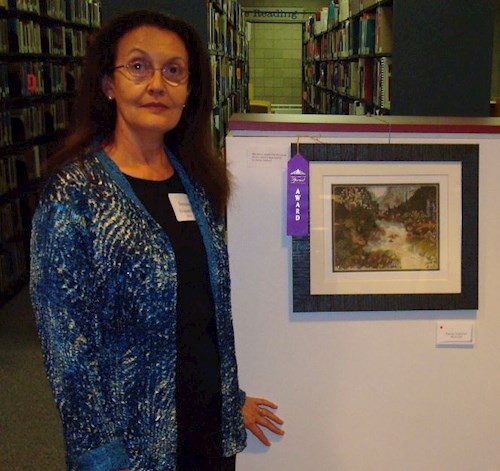 Photograph of a woman with long brown hair and glasses wearing a blue jacket standing next to a small, framed work of art that has a purple "award" ribbon attached to it 