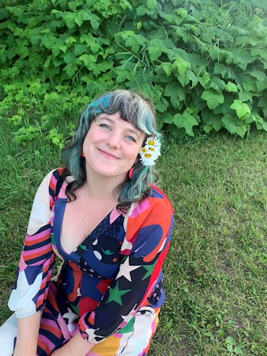 Image of a smiling woman with brown, blue and grey hair with white and yellow flowers next to her left ear. She is wearing a blue, red, purple and green dress. The background consists of green grass and green leaves.