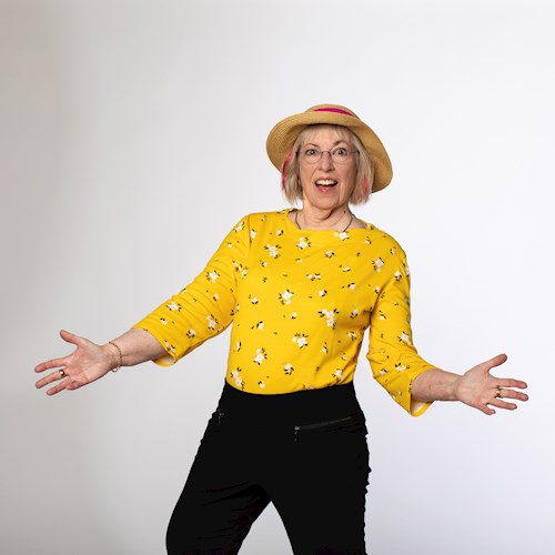 Image of a woman with short blonde hair wearing a straw-colored hat, bright yellow long sleeved shirt, and black pants. Her arms are extended outward in an open gesture of greeting. She is standing against a white background.
