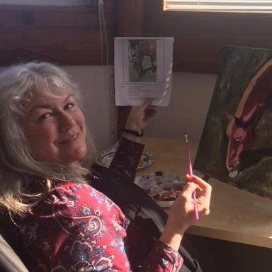 Image of a smiling woman with white hair holding up a reference photo in her left hand for a painting of a horse partially visible in the background. She holds a small paintbrush in her right hand. 