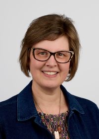 Headshot of a smiling woman with short brown hair, black glasses, and a denim jacket 