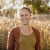 Photo of a smiling woman standing in front of a background of out-of-focus golden grasses. She has light brown hair and is wearing a rust-colored cardigan over a dark yellow top.