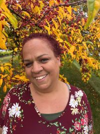 Photo of a smiling woman with short dark red hair, wearing a dark red top with white flowers. She is standing in front of a tree with yellow leaves and red berries.