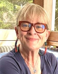 Photo of a smiling woman with blonde bangs, wearing red-framed glasses and a dark grey top.