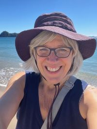 Photo of a smiling woman wearing glasses, a dark blue hat, and a sleeveless dark blue top. A beach is visible in the background. 