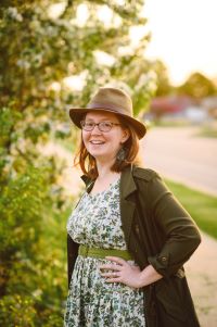 Photo of a smiling woman wearing a light green hat, glasses, a white a green dress, and a green jacket. Green foliage is visible in the background. 