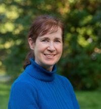 Person with brown hair pulled back smiles while wearing a bright blue turtleneck sweater, standing outdoors with blurred greenery in the background.