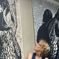 Person with curly blonde hair and glasses looking up at two large black-and-white rooster artworks on a wall. One rooster is highly detailed and prominent; the other is more abstract and shadowed.