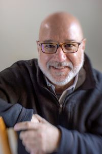 Person with a bald head, glasses, and a dark zip-up sweater over a collared shirt, smiling and leaning forward with one arm resting on a surface. Plain, softly blurred background.