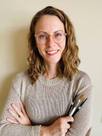 2.	Person with wavy light brown hair and glasses smiles while holding several paintbrushes, wearing a beige knit sweater against a plain background.