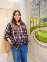 Person with brown hair wearing a plaid shirt and blue jeans, standing with hands in pockets between two large canvas artworks in an art studio. One canvas is abstract; the other depicts a road, grassy area, buildings, and a cloudy sky.