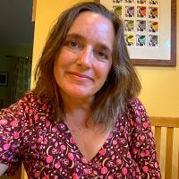 Person with shoulder-length brown hair smiling indoors, wearing a red floral top. Behind them is a yellow wall with framed artwork featuring a grid of colorful illustrations, in a dining area with a wooden chair and table.