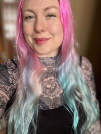 Person with long, wavy hair transitioning from pink to pastel blue, smiling slightly and wearing a black sleeveless top over a patterned long-sleeved shirt with a heart design. Soft indoor lighting and blurred background.