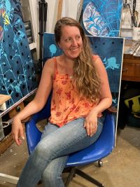 Person with long, light brown wavy hair wearing a red-orange floral sleeveless top and blue jeans, seated in a blue chair in an art studio. Background includes blue-toned botanical and abstract artworks and a workbench with art supplies.